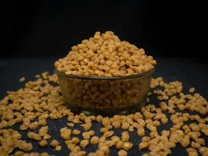 The image depicts a glass bowl of butterscotch nuts with a large amount of butterscotch nuts spilled around it.