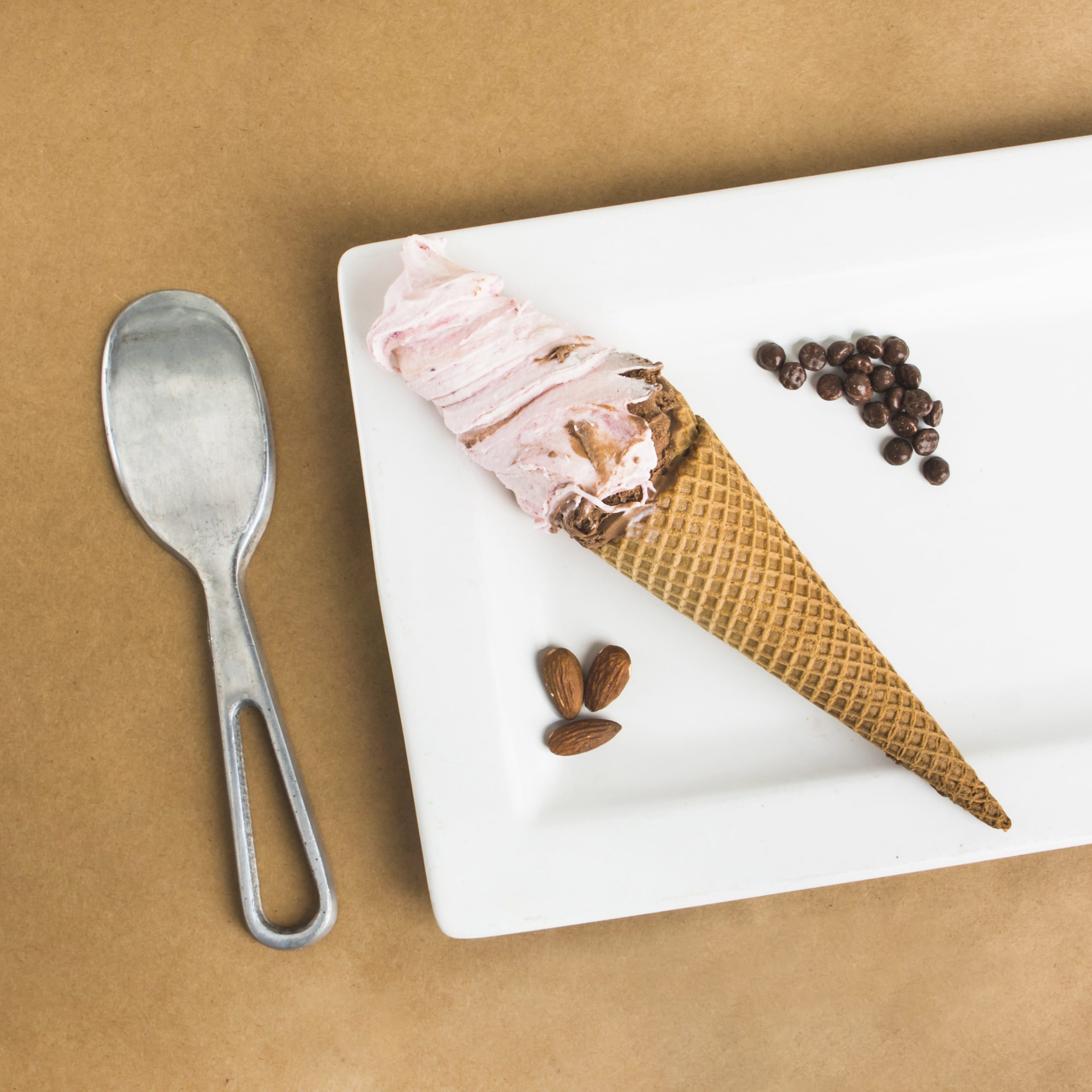 The pic shows a pink swirl of ice cream in a cone, sitting on a white rectangular plate. Three small almond halves are scattered on the plate to the left of the ice cream cone. A small amount of scattered chocolate chips are positioned to the right of the ice cream cone.